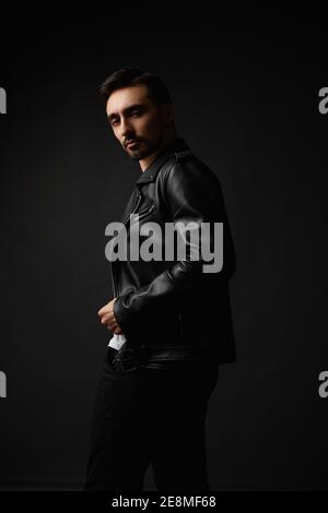 Young handsome man with unshaven face in black leather jacket posing in the dark background, isolate. Stylish bearded guy in a leather jacket Stock Photo