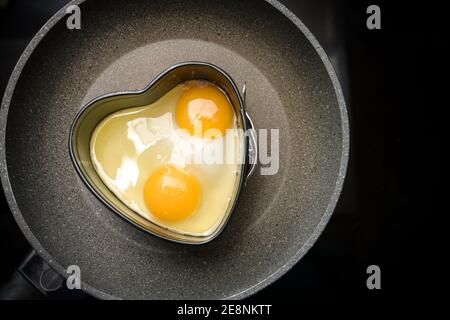 Two fried eggs in a heart shape on a frying pan on the black stove, preparation for a romantic valentines day breakfast, copy space, high angel view f Stock Photo