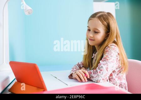 Child primary school age sit at desk with blank exercise book, hold pen and look at tablet screen, study at home during lockdown Stock Photo