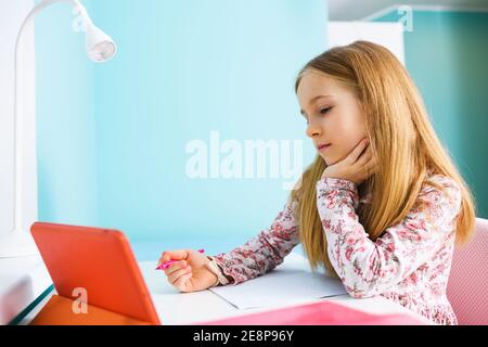 Child primary school age sit at desk with blank exercise book, hold pen and look at tablet screen, study at home during lockdown Stock Photo
