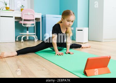 Primary school age sportive gymnast girl in black leotard stay in twine pose and look at tablet screen during online training at home Stock Photo