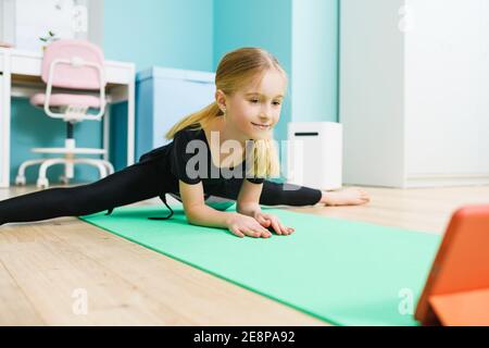 Primary school age sportive gymnast girl in black leotard stay in twine pose and look at tablet screen during online training at home