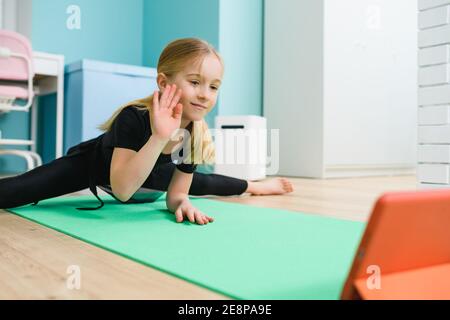 School girl gymnast in black leotard exercise at home mat, stay in twine pose during online lesson and wave hand to screen at isolation quarantine tim Stock Photo