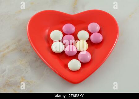 Pink, white and yellow Valentine's Day candy covered chocolate pieces on red heart shaped plate on table Stock Photo