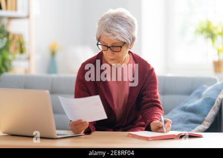 Elderly woman, sitting on the sofa with a paper receipt in her hands, are calculating expenses, managing the family budget. Stock Photo