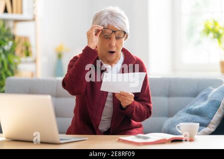 Elderly woman, sitting on the sofa with a paper receipt in her hands, are calculating expenses, managing the family budget. Stock Photo