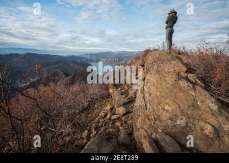 A female hiker pauses to stand on top of some rocks to overlook a valley and lake below in the California wilderness. Stock Photo