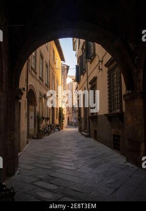 Beautiful street with Roman arches, yellow buildings, man riding a bike, in Lucca, Tuscany, Italy.  Sunny day, blue sky. Stock Photo