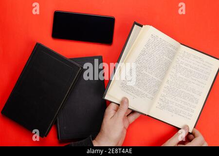A man's hand holds an open paper book on a red table. A black smartphone and two closed books lie side by side.  Flat lay Stock Photo