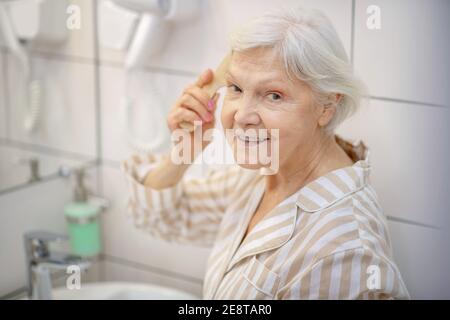 Gray-haired woman brushing her hair in the bathroom Stock Photo