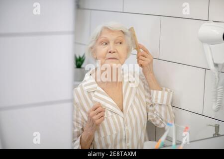 Gray-haired woman brushing her hair in the bathroom and feeling good Stock Photo