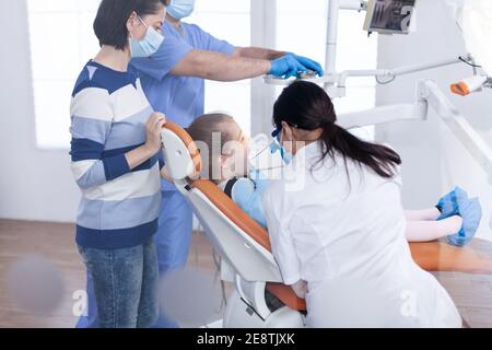 Kid getting cavity treatment from dentist sitting on dental chair wearing bib. Dentistry specialist during child cavity consultation in stomatology office using modern technology. Stock Photo