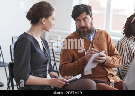 Group of people in audience at business conference focus on bearded man talking to woman while sitting on chairs Stock Photo