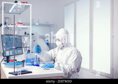 Microbiologist dressed in coverall using automatic pipette in healthcare laborator. Chemist in modern laboratory doing research using dispenser during global epidemic with covid-19. Stock Photo