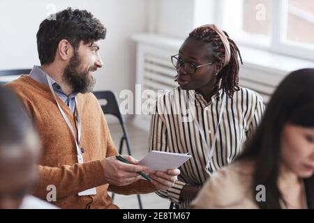 Multi-ethnic group of people in audience at business conference focus on bearded man talking to African-American woman while sitting on chairs Stock Photo