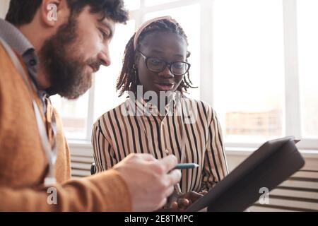 Portrait of young African-American woman talking to colleague and looking at tablet while discussing business project in office Stock Photo