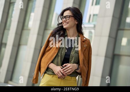 Portrait of a young happy woman holding smartphone, looking aside and smiling while walking on the city street on autumn day, stylish lady standing against blurred urban background. People lifestyle Stock Photo