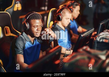 Young african guy, professional cybersport gamer wearing headphones talking by microphone with team member while playing online video game. eSport tournament concept Stock Photo