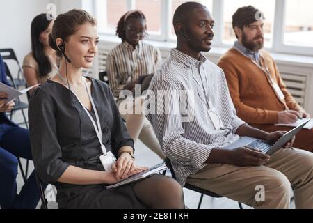 Multi-ethnic group of people sitting on chairs in audience at business conference or seminar, focus on young woman wearing headset in foreground Stock Photo