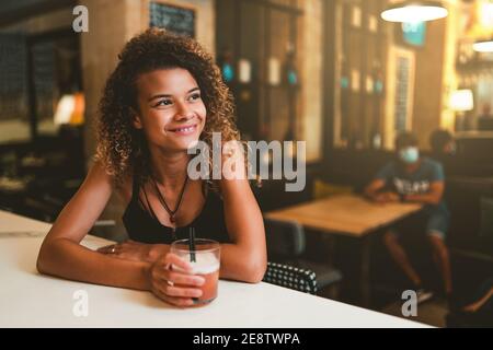 Happy young woman drinking at bar counter, man wearing a mask in the background Stock Photo
