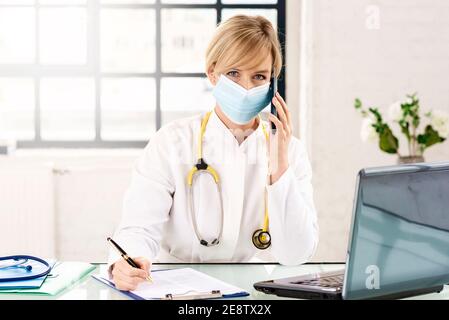 Portrat shot of smiling female doctor sitting at desk at using mobile phone while working in doctor’s office. Stock Photo