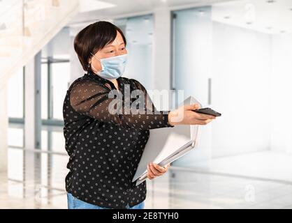 Asian woman using a surgical mask offering her phone while holding a folder at an office. Selective focus. Work and safety concept. Stock Photo