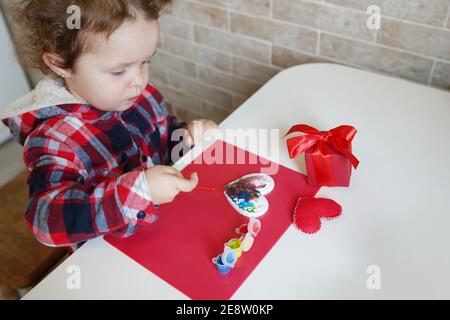 Little child girl decorates handmade Valentine heart by paints. Children's DIY, hobby concept, gift with your own hands. Valentine's Day decoration Stock Photo