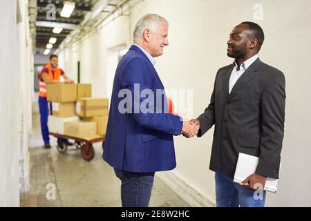 Two businessmen shaking hands in front of workers in the factory with packages on push trolleys Stock Photo