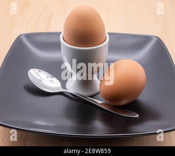 two eggs a spoon and an egg cup on a black plate isolated on a brown chopping board Stock Photo