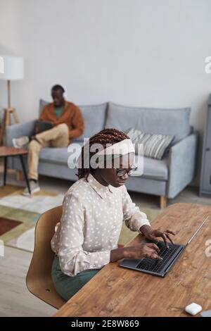 Vertical high angle portrait of young African-American woman using laptop at desk while working from home in cozy apartment interior Stock Photo