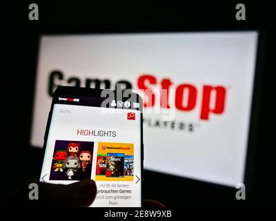 Person holding mobile phone with online shop of retail company GameStop Corp. on display with business logo in background. Focus on phone screen. Stock Photo