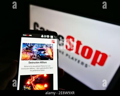 Person holding phone with online shop of retail company GameStop Corp. on display with logo in background. Focus on center of cellphone screen. Stock Photo