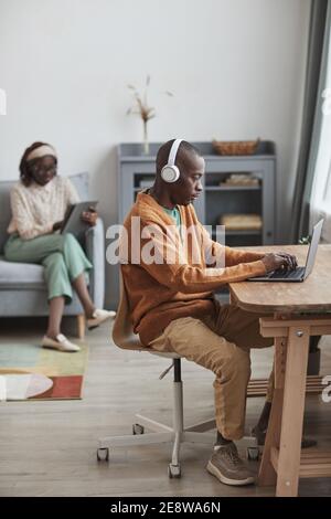 Vertical portrait of contemporary couple using internet devices at home, focus on African-American man wearing headphones and typing at desk in foregr Stock Photo