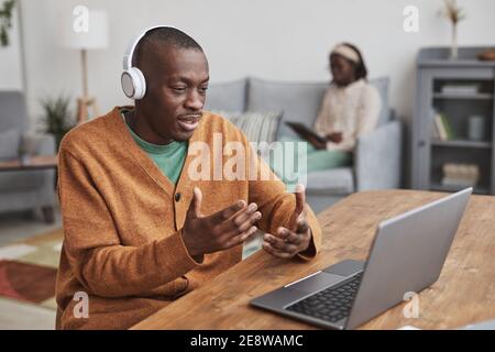 Portrait of African-American man using laptop for video chat and wearing headphones while sitting at desk while in minimal home interior, copy space Stock Photo