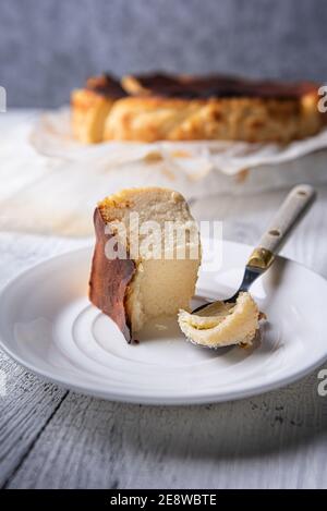 Bitten portion of basque burnt cheesecake on white plate with vintage spoon on side. Unfocused whole cheesecake at background. Homemade spanish desser Stock Photo