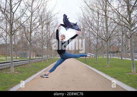 Handsome young man wearing make up and headphones, doing a ballet jump in a park path. Non binary androgynous guy. Stock Photo