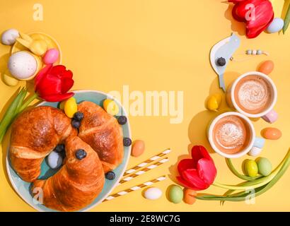 Spring breakfast with croissants and berries. Two Espresso coffee in bright cups, Easter chocolate eggs, pink bunny ears and red tulips on a yellow background. Copy space. Flat lay Stock Photo