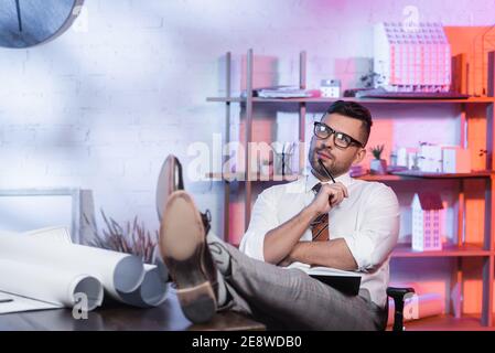 thoughtful architect looking away while sitting with legs on desk Stock Photo