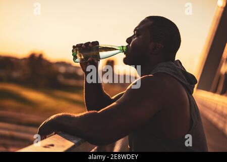 Portrait of young african-american man in sports clothing who is drinking water after exercise. He is exercising to reduce his body weight. Stock Photo