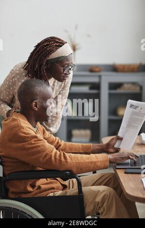 Vertical side view portrait of African-American man using wheelchair working from home with wife looking over his shoulder, copy space Stock Photo