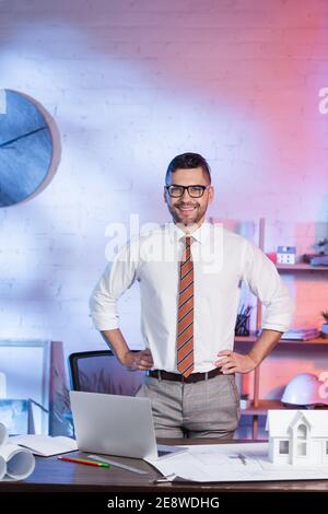 happy architect standing with hands on hips near laptop and house model on desk Stock Photo
