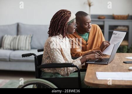 Side view portrait of young African-American woman using wheelchair working from home with husband helping her copy space Stock Photo