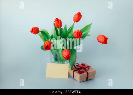 Red tulips in glass vase and gift in a box on blue background. Valentines or Women's day concept. Copy space. Stock Photo