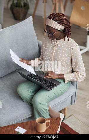 Vertical high angle portrait of young African-American woman wearing glasses and using laptop while sitting on sofa and working from home Stock Photo