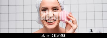 cheerful woman in towel on head using cleansing silicone brush in bathroom, banner Stock Photo