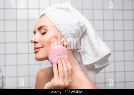 pleased woman in towel on head using cleansing silicone brush in bathroom Stock Photo
