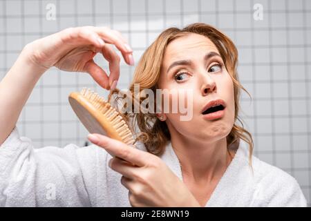 dissatisfied woman in bathrobe brushing hair in bathroom Stock Photo