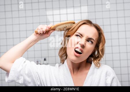 displeased woman in white bathrobe brushing hair in bathroom Stock Photo
