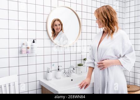 joyful woman in bathrobe smiling while looking at mirror in bathroom Stock Photo
