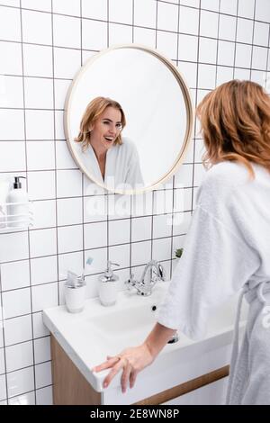 cheerful woman in bathrobe smiling while looking at mirror in bathroom Stock Photo
